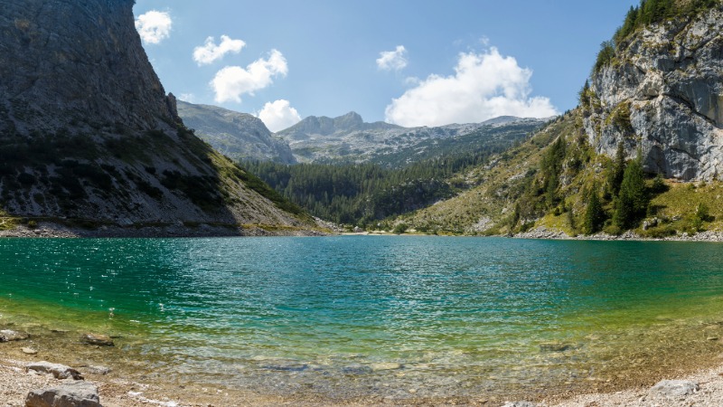 Pogled na kristalno čisto Krnsko jezero, obdano z gozdovi in strmimi gorami Julijskih Alp v sončnem jesenskem dnevu.