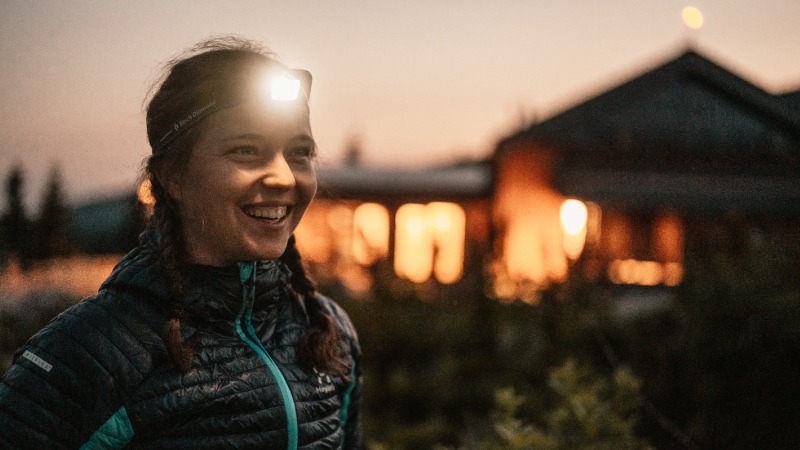 Nasmejana pohodnica v jesenski jakni, z delujočo naglavno svetilko na čelu, v mraku pred gorsko kočo, kar poudarja pomen osvetlitve.