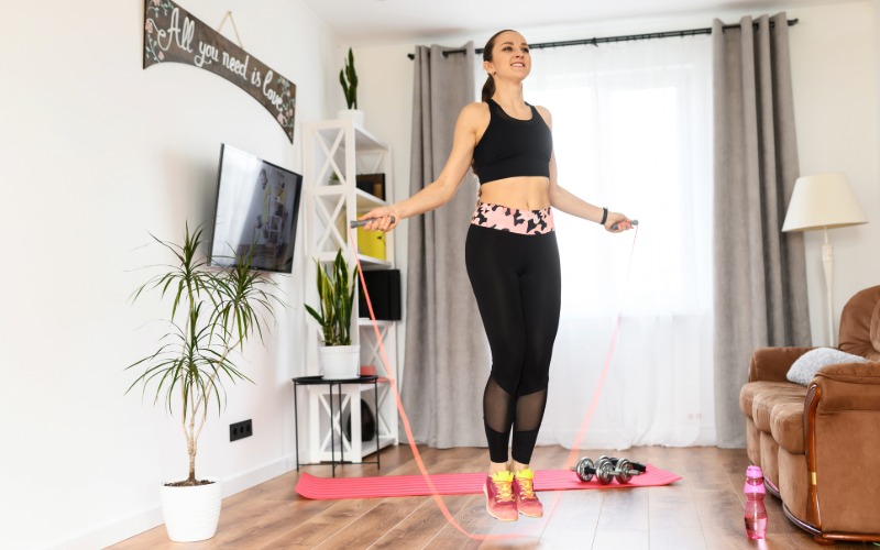 Ženska v športnih oblačilih izvaja vajo s kolebnico v dnevni sobi kot del HIIT treninga doma.