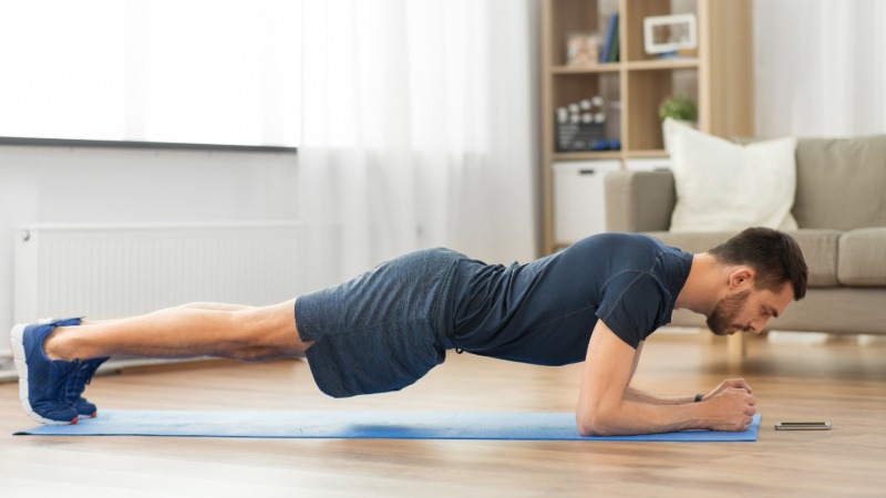 Tekač izvaja vajo deska (plank) na podlogi v dnevni sobi za krepitev jedra, kar je pomemben del treninga za moč.