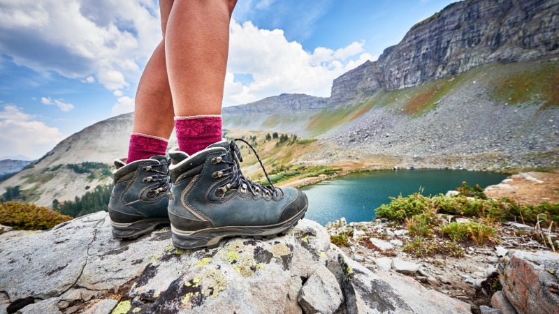 Pohodne noge v udobnih, dobro uhojenih gojzarjih stojijo na skali ob gorskem jezeru, simbolizirajoč pripravo na pohod.