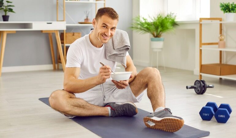 Nasmejan moški sedi na vadbeni podlogi po končanem treningu doma in uživa v obroku, bogatem z beljakovinami in ogljikovimi hidrati, za optimalno regeneracijo.