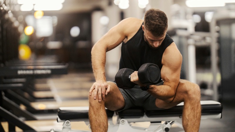 Moški v fitnesu počasi in kontrolirano izvaja koncentracijski upogib komolca z ročko, osredotočen na občutek v bicepsu.