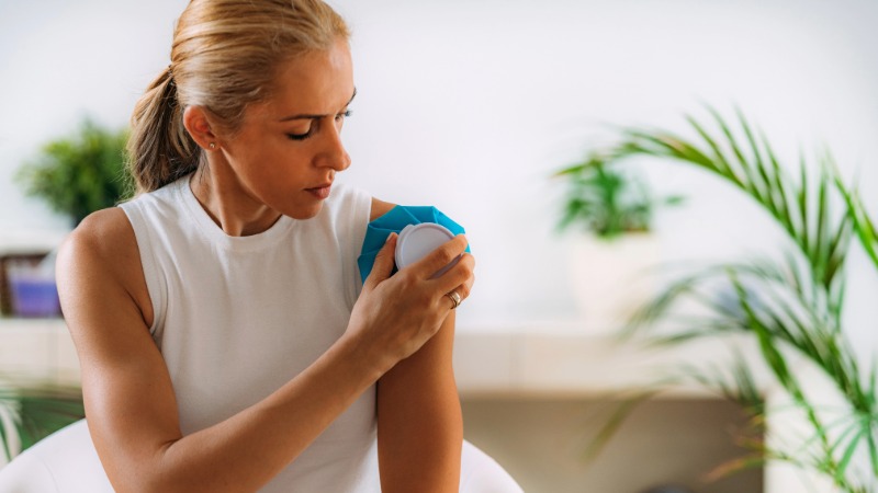 Ženska si z modrim hladnim obkladkom hladi ramo, kar je primer prve pomoči pri akutni poškodbi za zmanjšanje vnetja in otekline.
