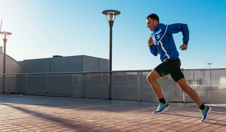 Moški tekač v modri tekaški jakni in črnih kratkih hlačah teče po tlakovanem pločniku na sončen dan. Njegovo telo je v gibanju, kolena dvignjena, stopala pa hitro prestopajo. V ozadju so vidne sodobne stavbe in ulične svetilke, nebo je jasno modro