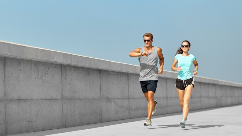 Moški in ženska v športnih oblačilih tečeta skupaj ob betonskem zidu. Moški med tekom preverja podatke na svoji športni uri, kar ponazarja merjenje tekaške metrike, kot je kadenca