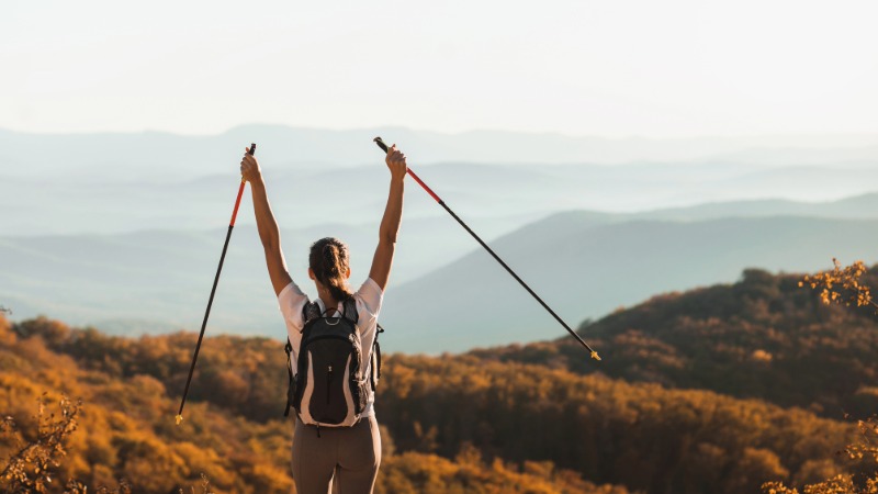 Ženska stoji na razgledni točki v gorah, z dvignjenimi pohodniškimi palicami, kar simbolizira svobodo in užitek v nordijski hoji kot alternativi za trening v coni 2.