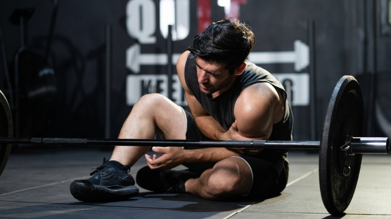 Moški sedi na tleh v fitnesu in se z bolečino drži za ramo, kar ponazarja primer akutne poškodbe rame med vadbo z utežmi.