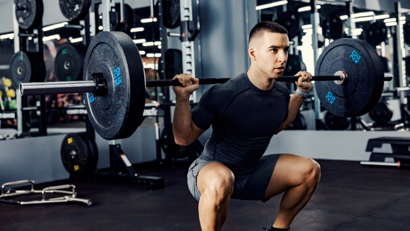 Moški izvaja počep z naloženo palico v fitnesu, vajo za krepitev povezave med možgani in mišicami nog ter zadnjice.