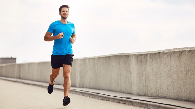 Nasmejan tekač v modri majici teče z vzravnano držo in pogledom, usmerjenim naprej, kar ponazarja pravilno tehniko teka.