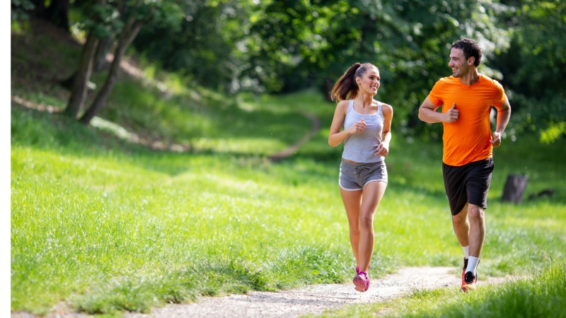 Moški in ženska tečeta skupaj po gozdni poti, se sproščeno pogovarjata in kažeta primer "pogovorne cone" pri treningu v coni 2.