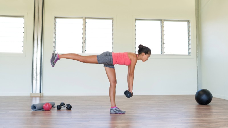 Prikaz izvedbe enonožnega romunskega mrtvega dviga (RDL) z utežjo v roki. Ženska stoji na eni nogi, trup je vzporeden s tlemi, hrbet pa raven.