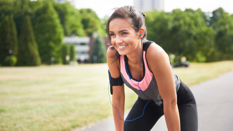 Nasmejana tekačica z brezžičnimi slušalkami uživa v lahkotnem teku v parku, kar ponazarja zmanjšanje stresa in izboljšanje počutja z vadbo v coni 2.