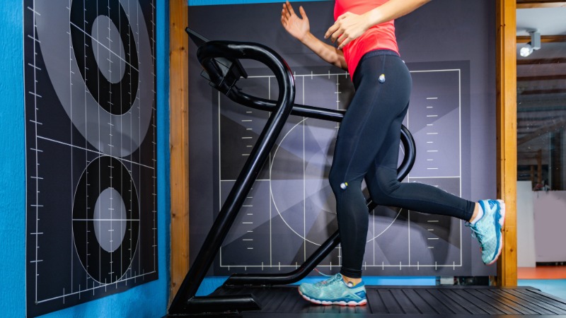 Tekačica na tekaški stezi v laboratoriju za analizo teka, kjer se meri kadenca in tehnika koraka.