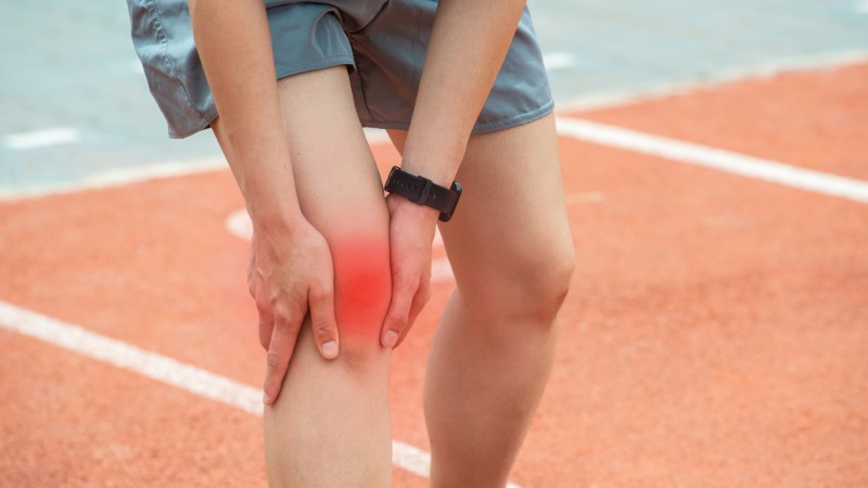 Tekačica se na stezi drži za koleno, z rdečim poudarkom na sprednjem delu, ki ponazarja lokacijo bolečine pri tekaškem kolenu.