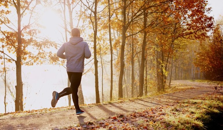 Tekač v lahki jesenski opremi teče po sončni gozdni poti, polni zlatih listov, kar simbolizira umirjenost in uživanje v teku v coni 2.