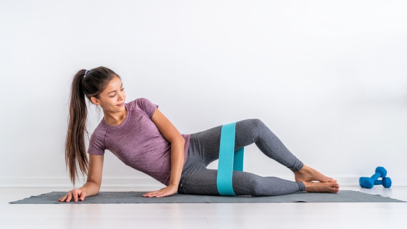 Prikaz vaje "školjka" (clamshell) z modro elastiko okoli stegen za krepitev stranskih mišic kolka.