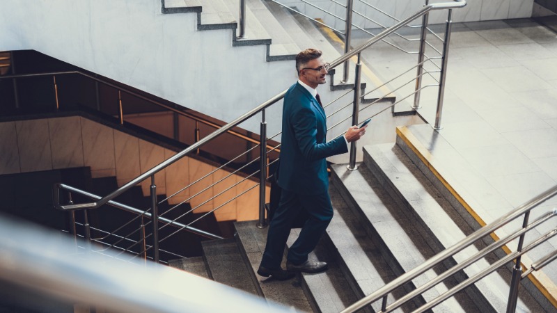 Moški v poslovni obleki hodi po stopnicah v moderni stavbi in gleda na svoj pametni telefon, s čimer ponazarja nabiranje korakov med delovnim dnem.