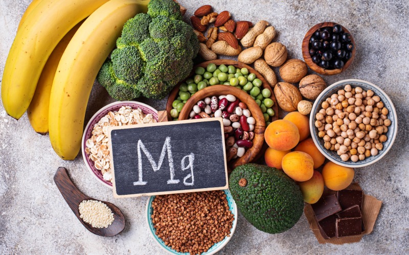 Raznolika in barvita zbirka živil, ki podpirajo uravnavanje stresa, razporejena na svetli podlagi. Vključuje banane, avokado, brokoli, oreščke, semena in stročnice. V sredini je majhna tabla s kemijskim simbolom za magnezij, Mg.