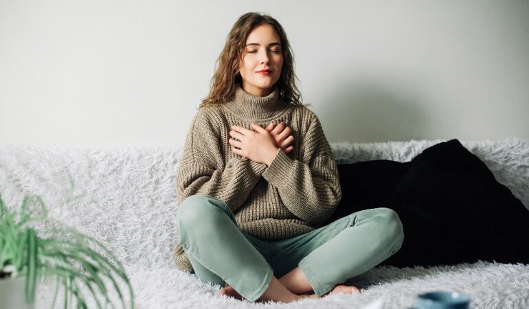 Ženska sedi v lotosovem položaju na belem kosmatem kavču, z zaprtimi očmi in rokama položenima na prsi. Na sebi ima debel bež pulover in svetlo zelene hlače. V ozadju je bela stena, ob strani pa je viden del zelene sobne rastline.