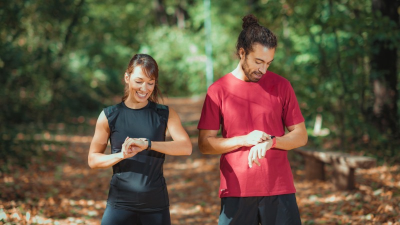 Moški in ženska v športnih oblačilih pregledujeta podatke o aktivnosti na svojih pametnih urah med sprehodom v naravi.