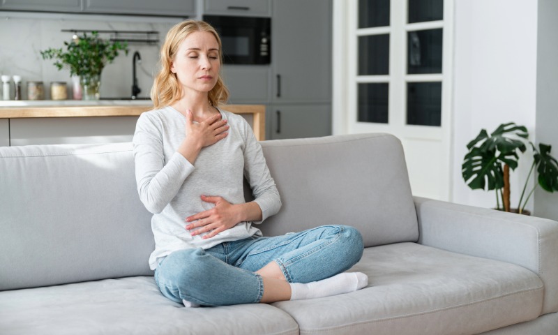 Ženska sedi s prekrižanimi nogami na sivem kavču z zaprtimi očmi. Eno roko ima položeno na prsi, drugo pa na trebuh, medtem ko mirno izvaja vajo zavestnega dihanja v domačem okolju.