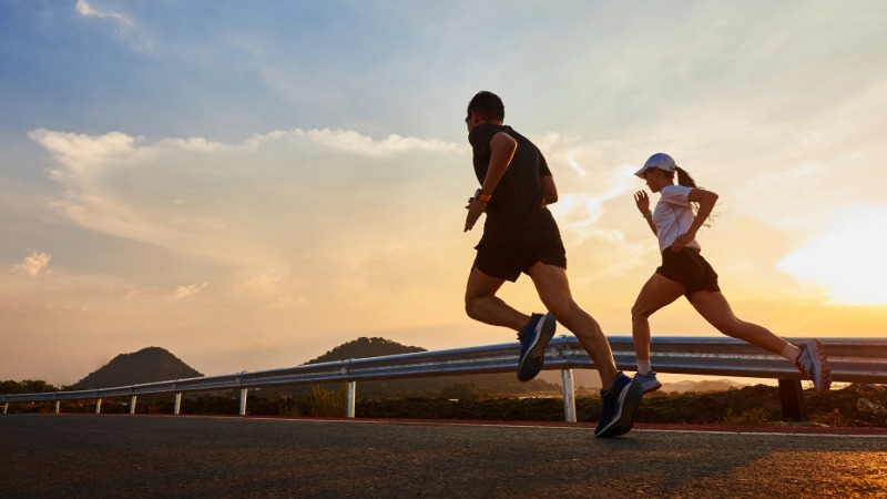 Dva tekača med intenzivnim treningom na cesti ob sončnem zahodu, kar ponazarja fizični stres, ki vpliva na variabilnost srčnega utripa.