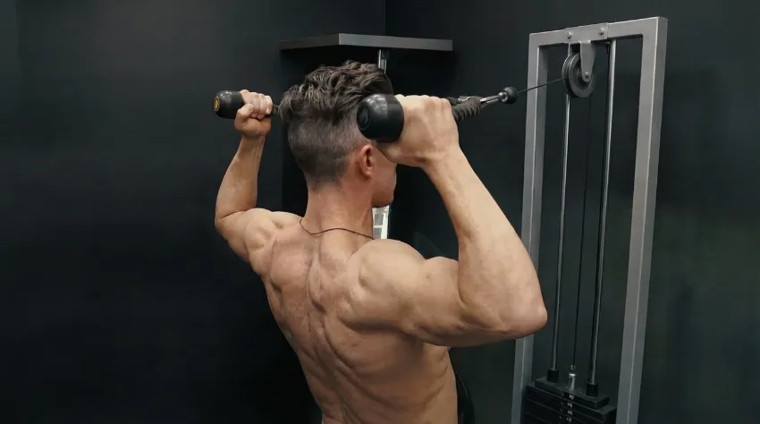 Moški na fitnes napravi izvaja končni položaj vaje Face Pulls. Njegovi komolci so visoko v ravnini z rameni, roke so aktivno rotirane navzven tako, da so pesti za ravnino komolcev, lopatice pa so stisnjene skupaj, kar spominja na pozo "dvojni biceps" od zadaj.