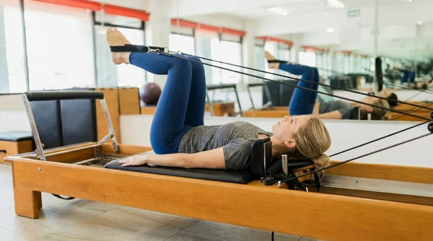 Ženska v studiu izvaja nadzorovano vajo na lesenem pilates reformerju z uporabo trakov za noge, kar omogoča varno krepitev trupa in razbremenitev hrbtenice.