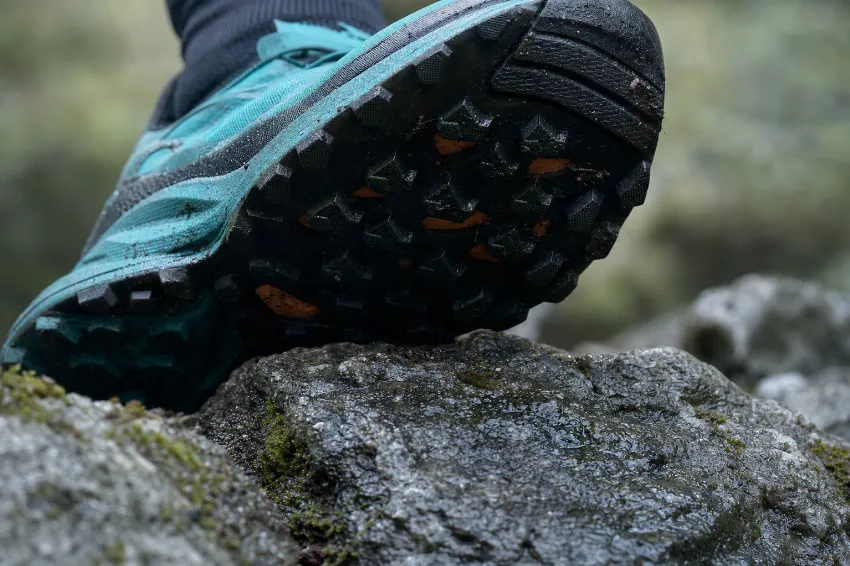Bližnji posnetek podplata trail superge, ki se trdno oprijema mokre skale na gorski poti.