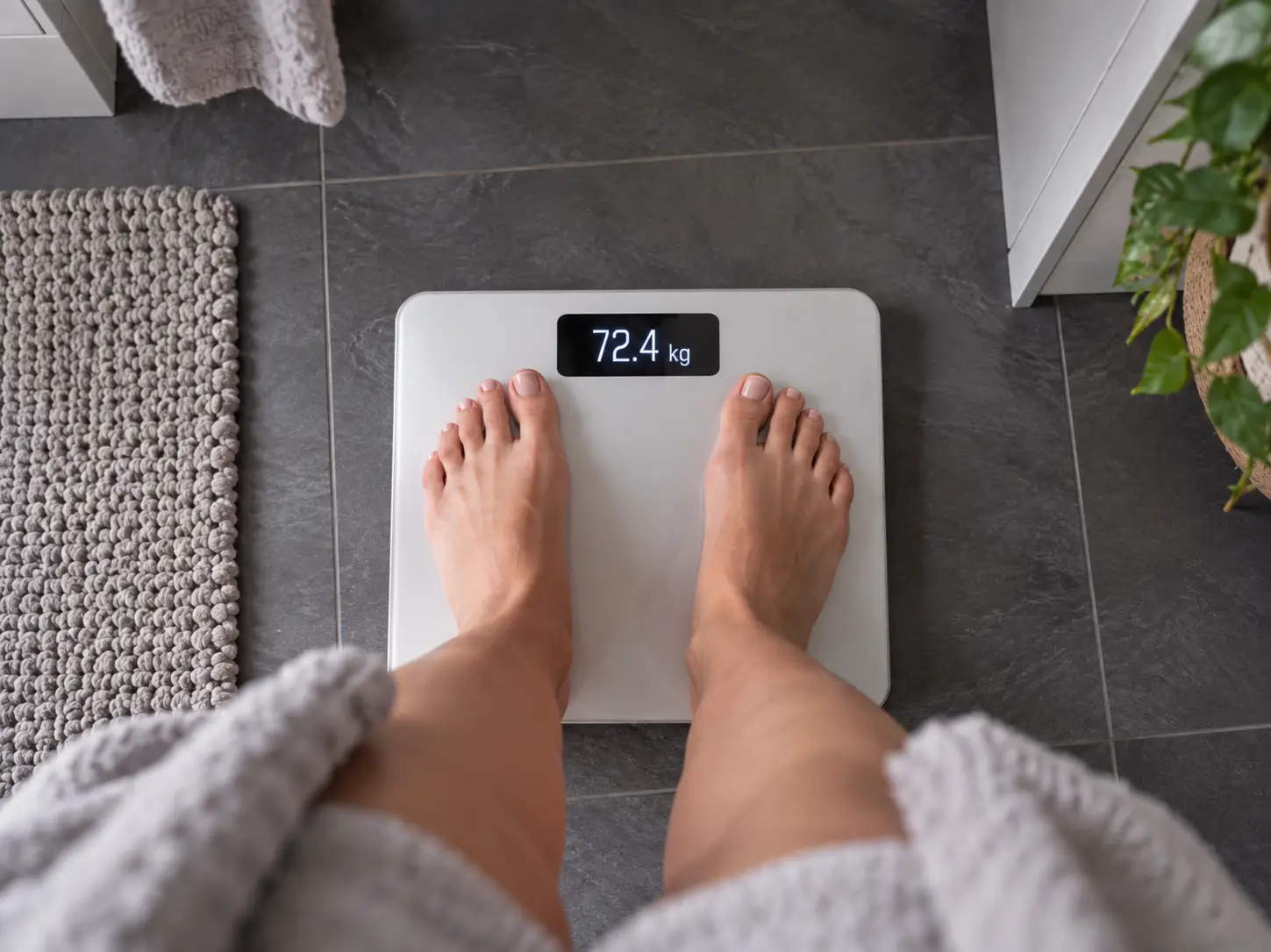Oseba med jutranjim tehtanjem na digitalni tehtnici, ki prikazuje 72,4 kg